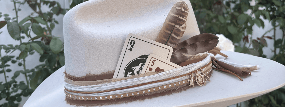 A cream-colored felt cowboy hat decorated with brown feathers, playing cards, and a decorative band with gold studs and a small gold star charm, set against a blurred green garden background at a custom hat bar event.
