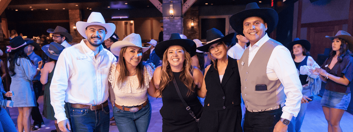 A group of five people posing together in a dimly lit indoor event space, dressed in western-style attire including cowboy hats, vests, and jeans at a custom hat bar event.
