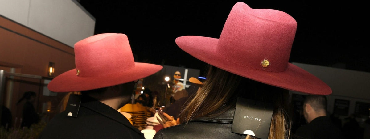 Two people wearing red hats with gold pins, one in a black coat and the other in a leather jacket, at night at a custom hat bar event.
