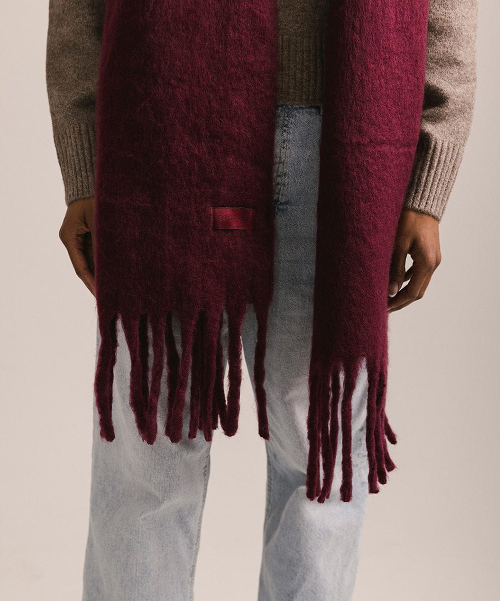Maroon scarf with fringe held by a person wearing a striped sweater and jeans on a beige background #color_merlot