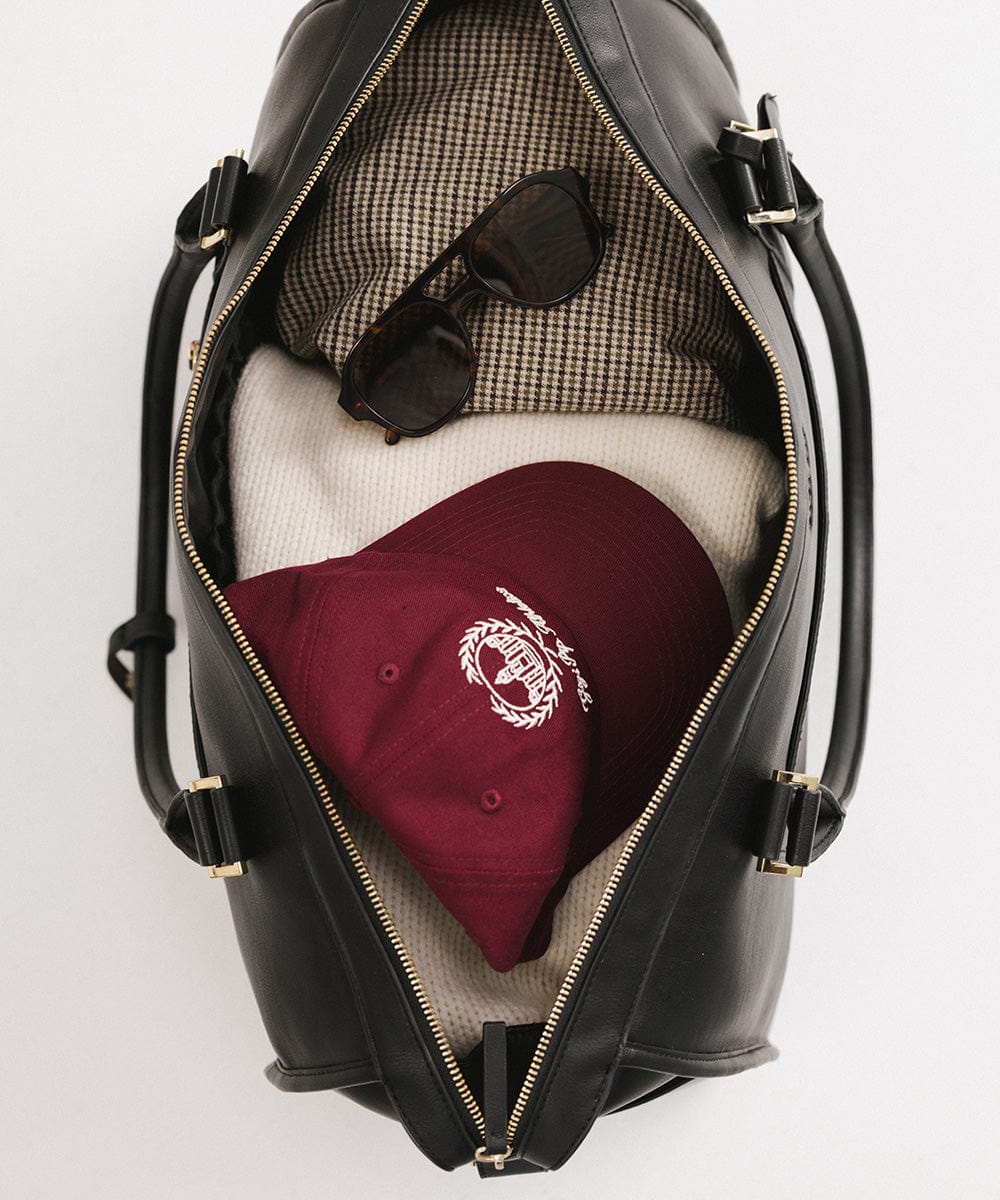 A black vegan leather weekender bag with a cap, sunglasses and clothes inside it against a plain background #color_black