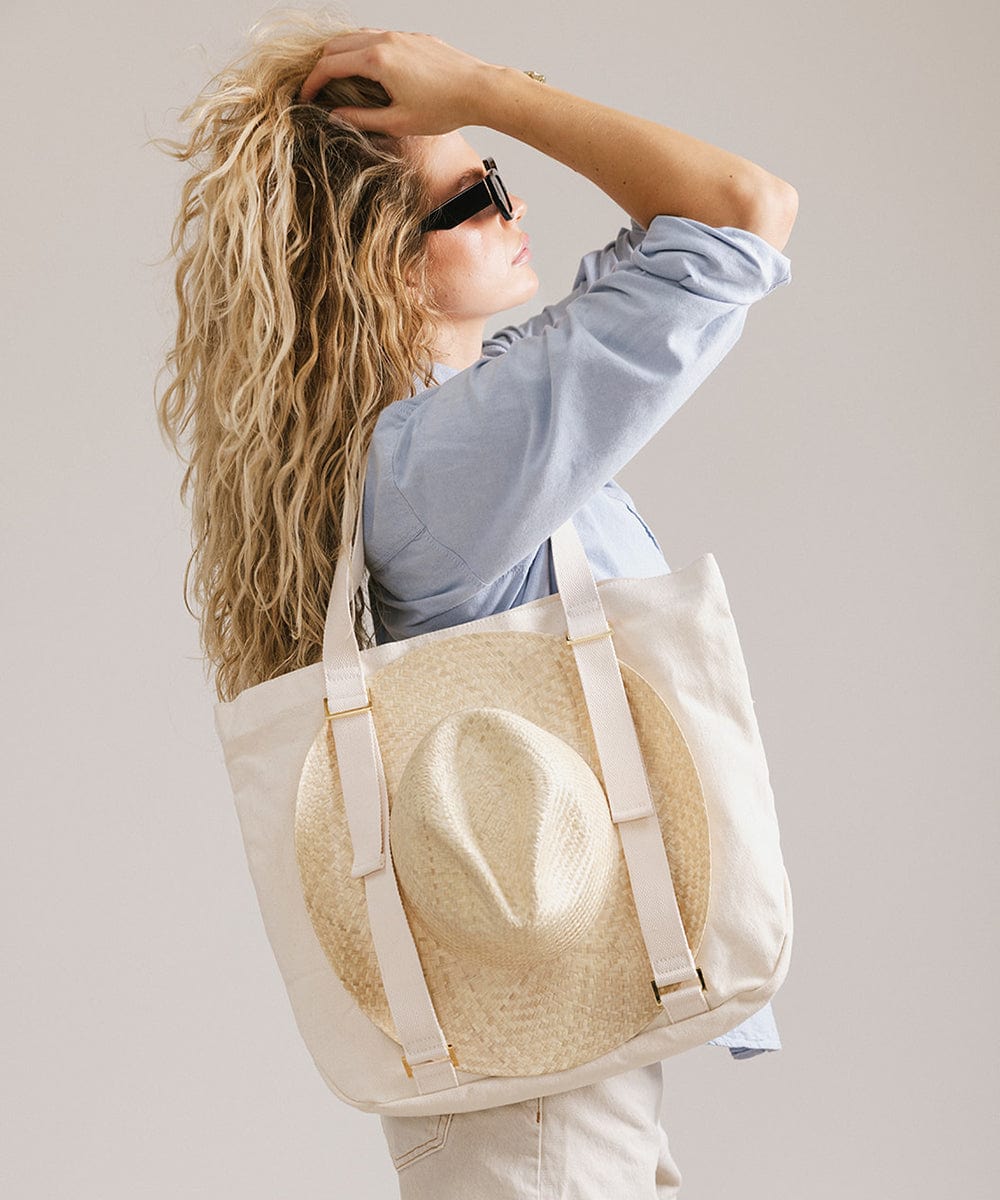 Woman wearing sunglasses, a blue shirt and white shorts, carrying a hat on a natural colored canvas tote bag, facing sideways against a plain background #color_natural