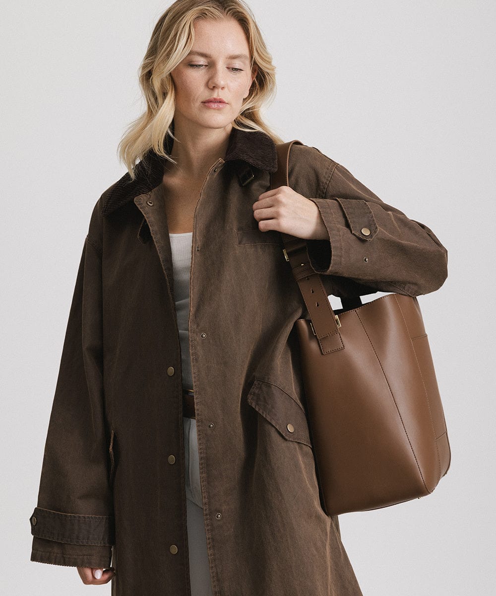 Woman wearing a brown coat carrying a chocolate colored Dia everyday leather tote bag against a plain background #color_chocolate