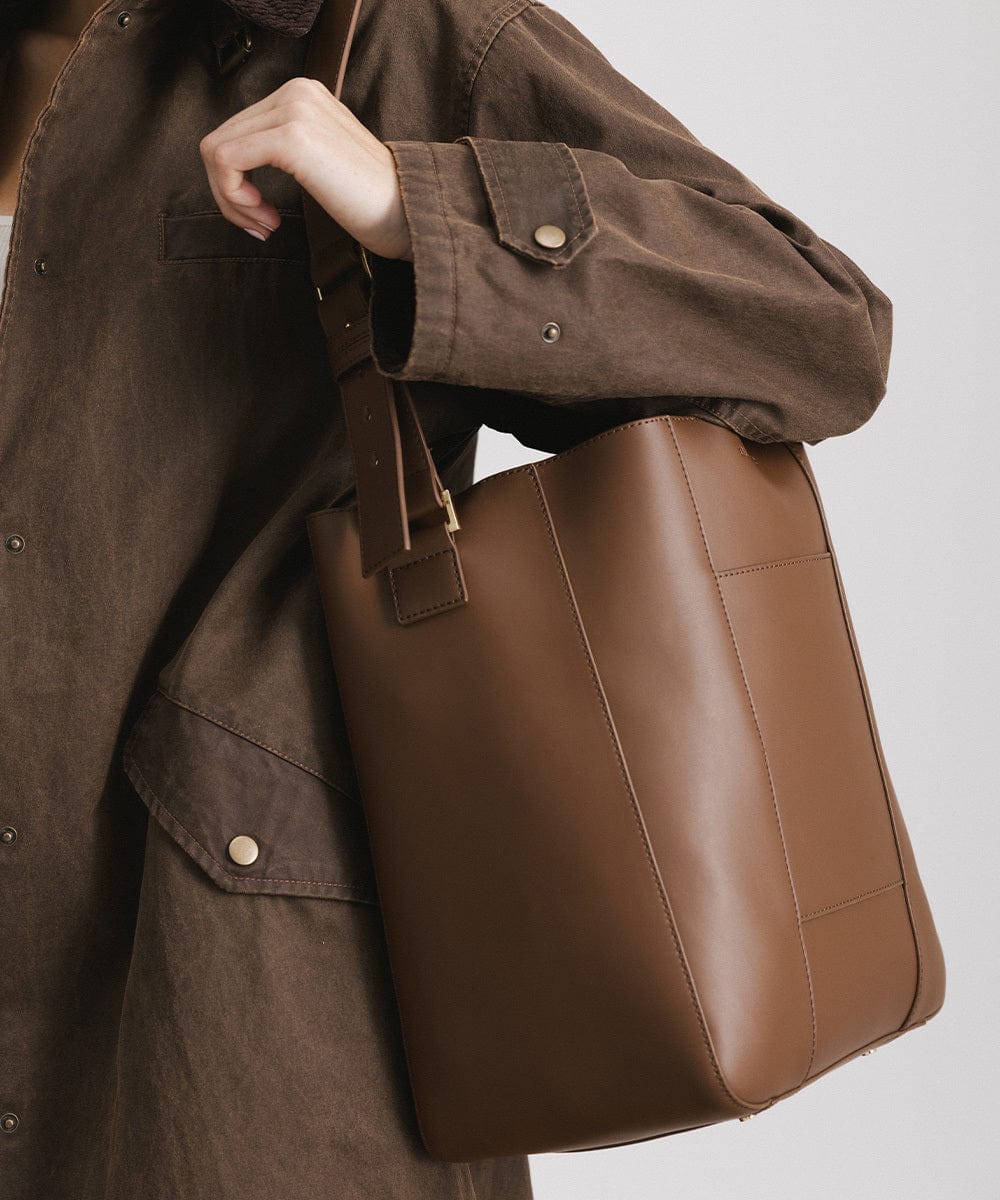 A person wearing a brown coat carrying a chocolate colored Dia Everyday Leather Tote Bag against a plain background #color_chocolate