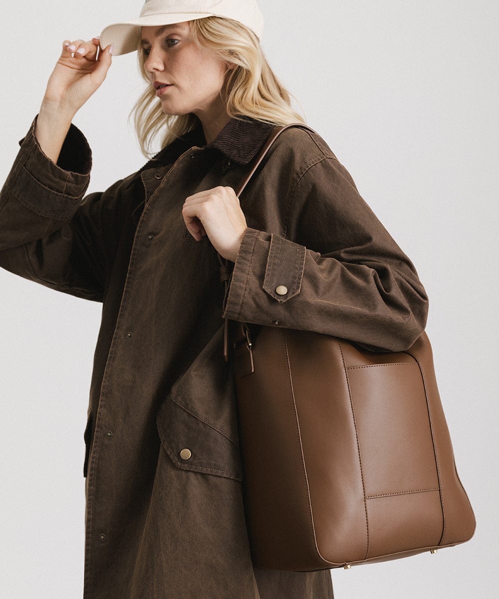 Woman wearing a white cap and a brown coat, carrying a chocolate colored Dia everyday leather tote bag, posing sideways against a plain background #color_chocolate