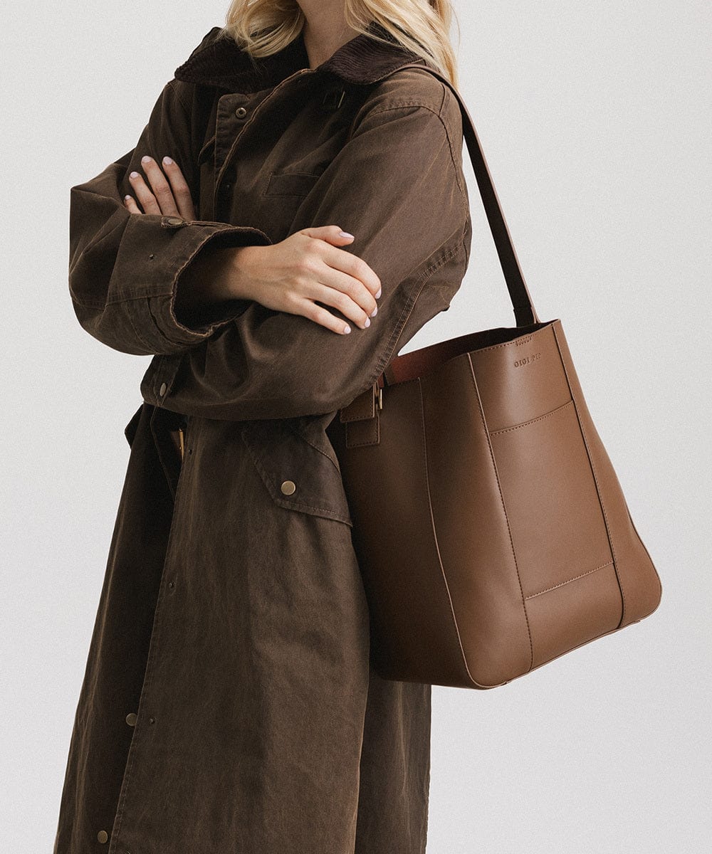 Person wearing a brown coat carrying a chocolate colored Dia everyday leather tote bag, posing against a plain background #color_chocolate