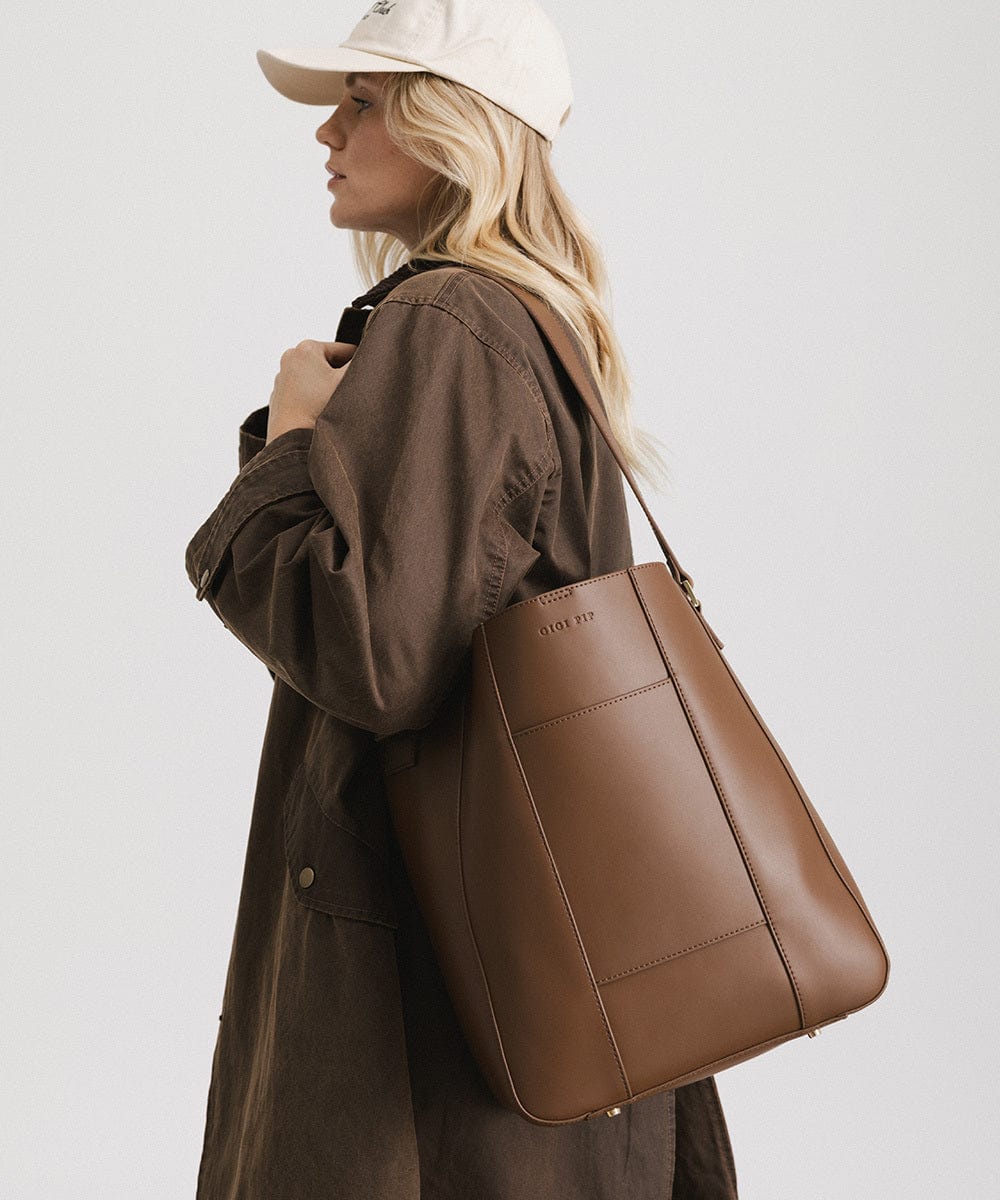 Woman wearing a brown coat carrying a chocolate colored Dia everyday leather tote bag, facing sideways against a plain background #color_chocolate