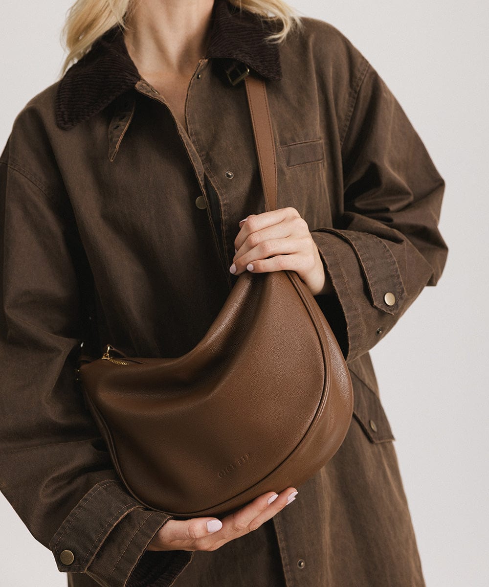 Person wearing a brown shirt carrying a chocolate colored genuine leather slouchy bag against a plain background #color_chocolate