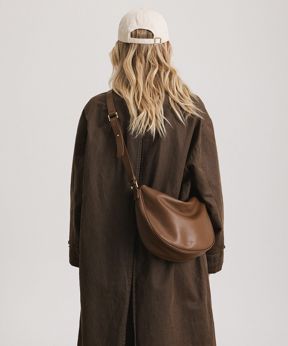 Person wearing a brown coat and a white cap, carrying a chocolate colored genuine leather slouchy bag against a plain background #color_chocolate