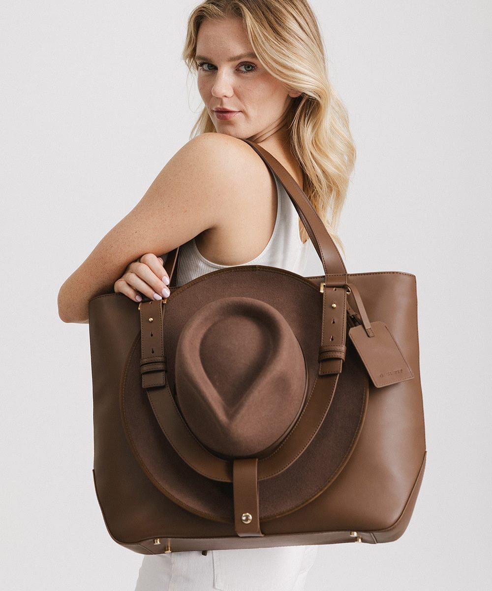 Woman carrying a brown Hayes Hat Carrying Tote Bag against a plain background #color_chocolate