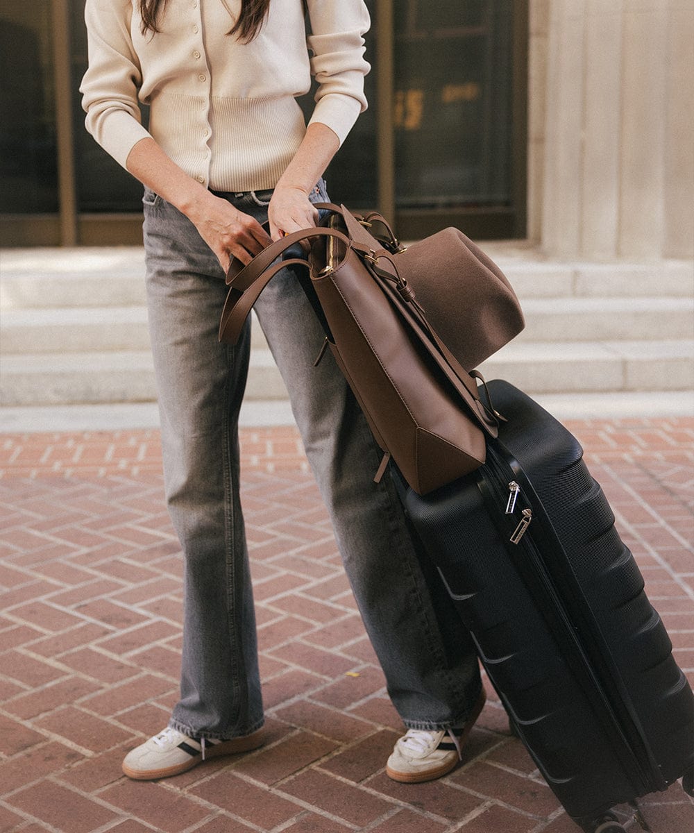 Person holding a brown leather bag and black suitcase on a brick pavement. #color_chocolate