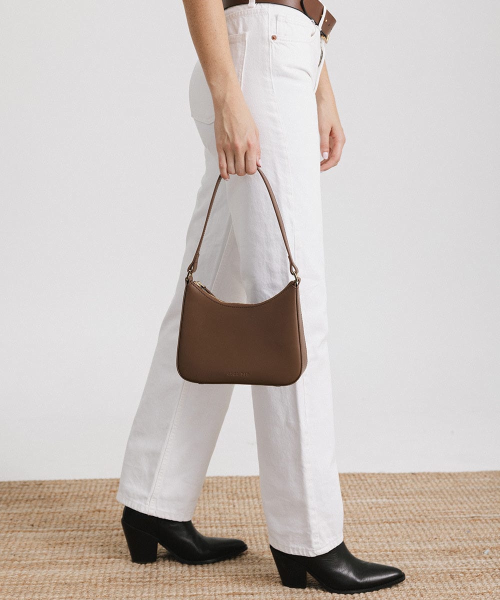 Person wearing a brown belt, white pants, and black shoes, carrying a chocolate colored genuine leather handbag against a plain background #color_chocolate