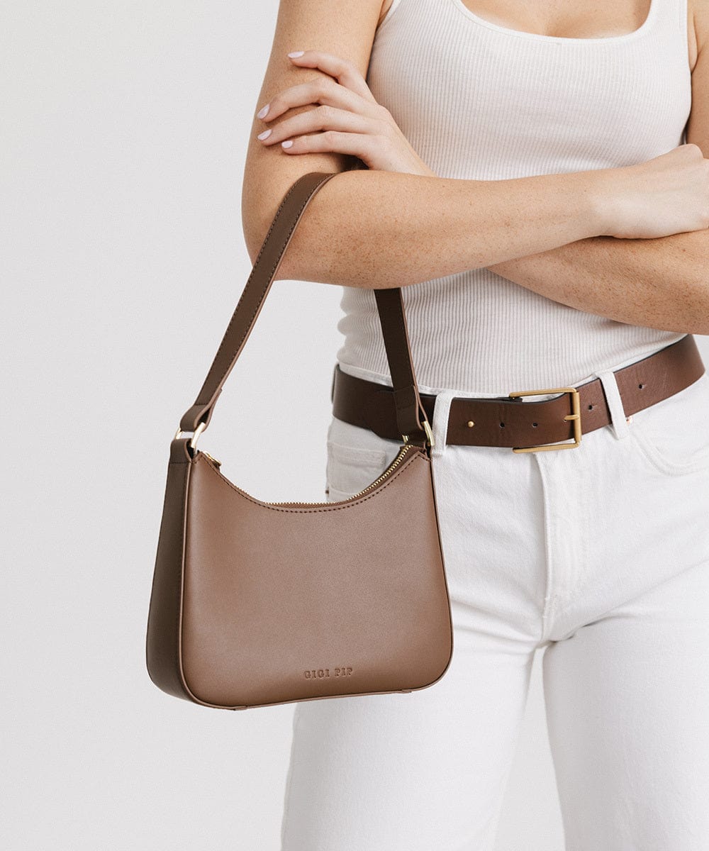 Person wearing a white top, brown belt, and white pants, carrying a chocolate colored genuine leather handbag against a plain background #color_chocolate