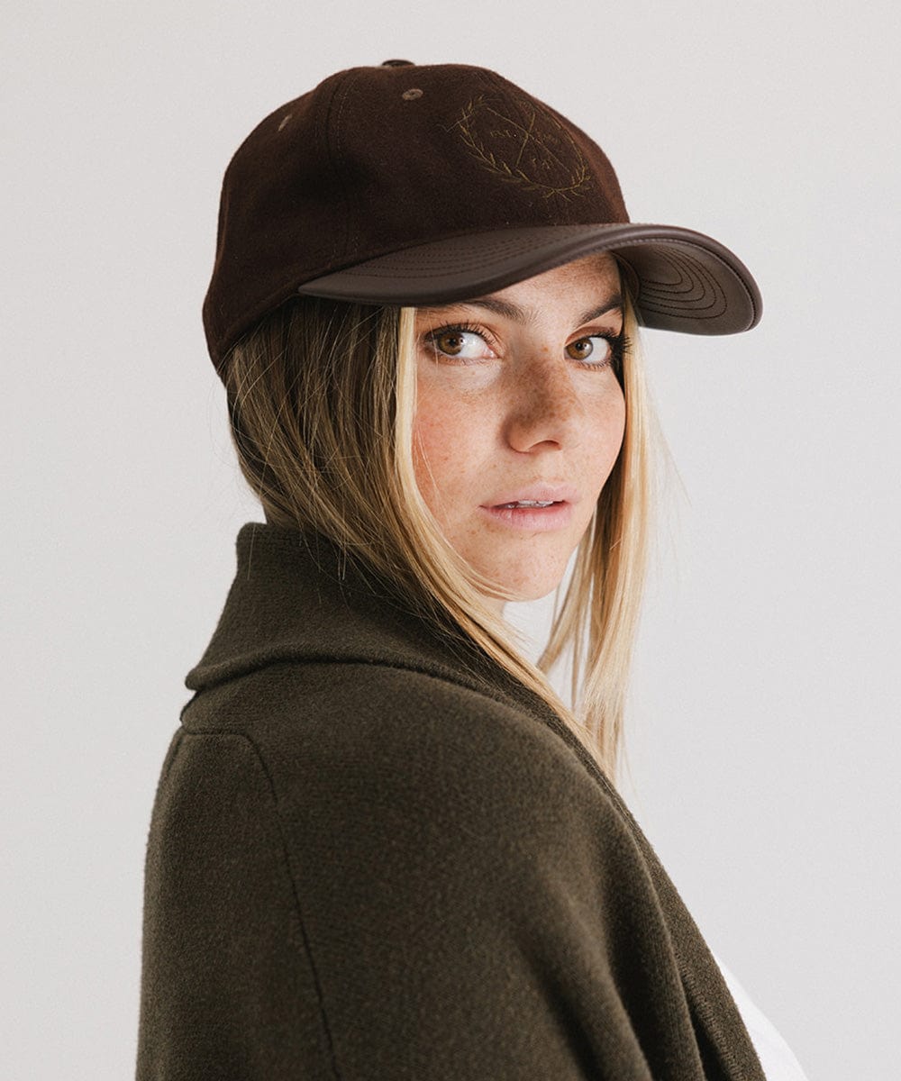 Woman wearing a brown cap with a logo and a white t-shirt with a brown coat, posing sideways against a plain background #color_brown