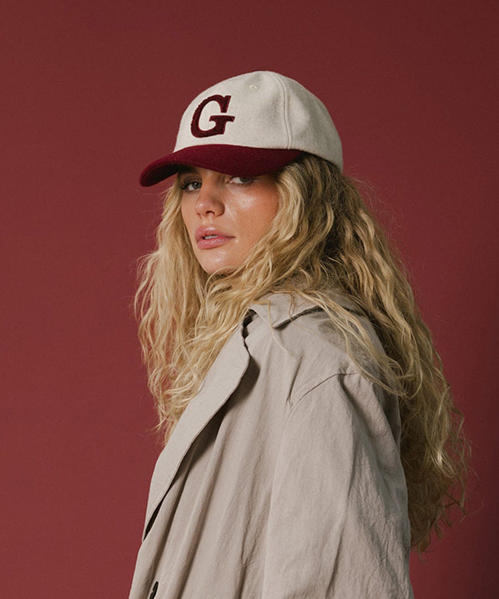 Woman wearing a white and burgundy baseball cap with the letter 'G' and a beige shirt against a red background #color_burgundy