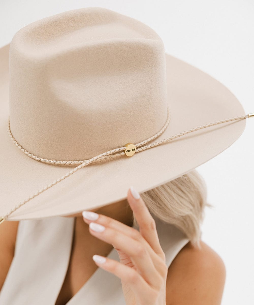 Close up image of a woman wearing a cream cowboy hat with a cream braided hat band and a white top against a plain background #color_cream