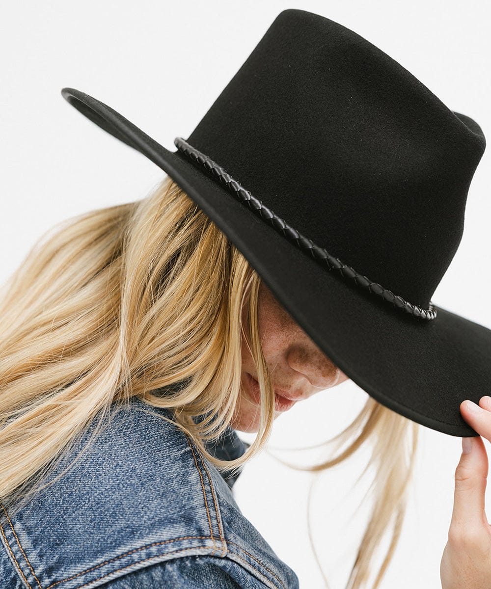 Woman wearing a black hat with a black braided bolo hat band and a denim jacket, posing against a plain background #color_black
