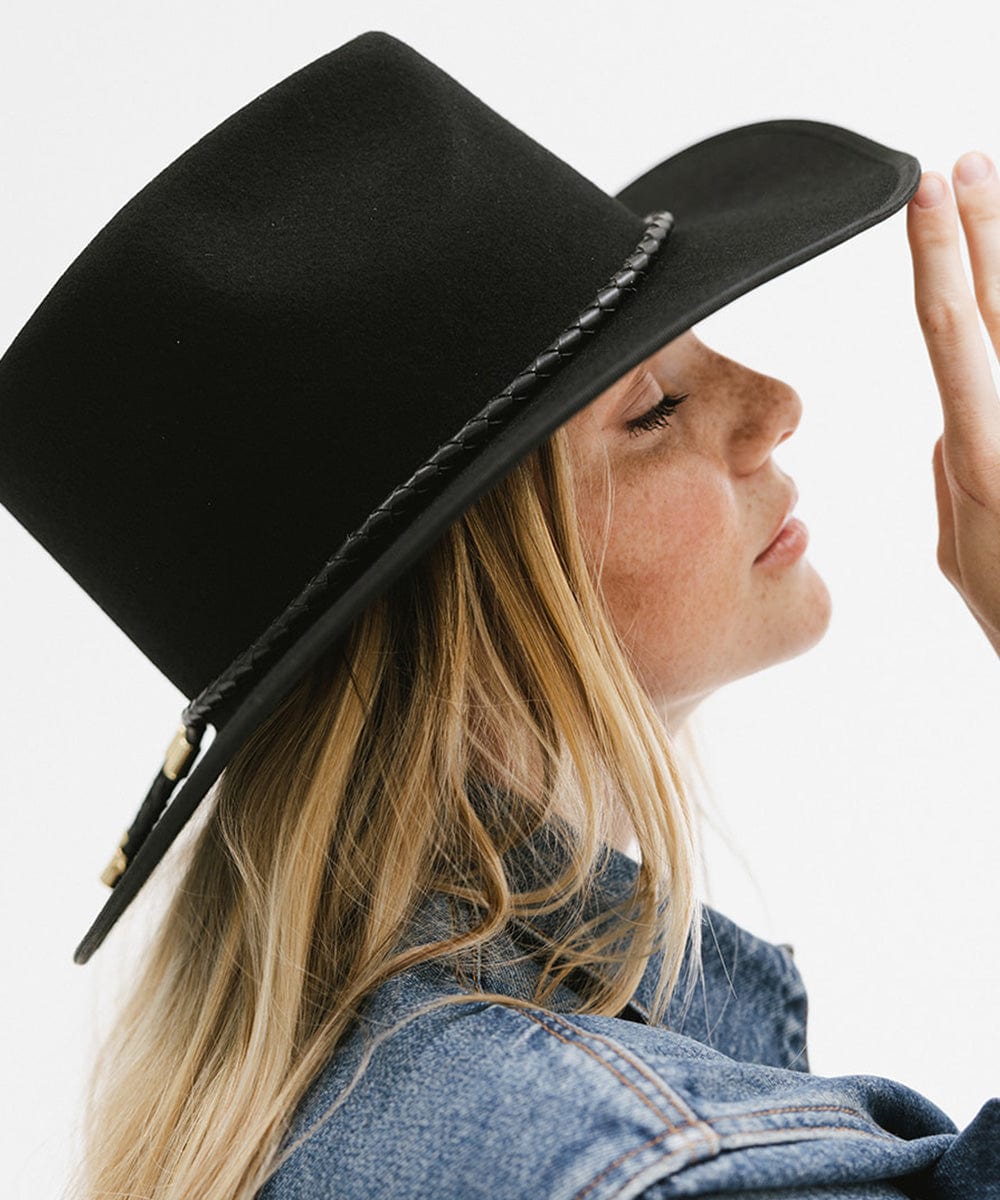 Woman wearing a black hat with a black braided bolo hat band and a denim jacket, facing sideways against a plain background #color_black