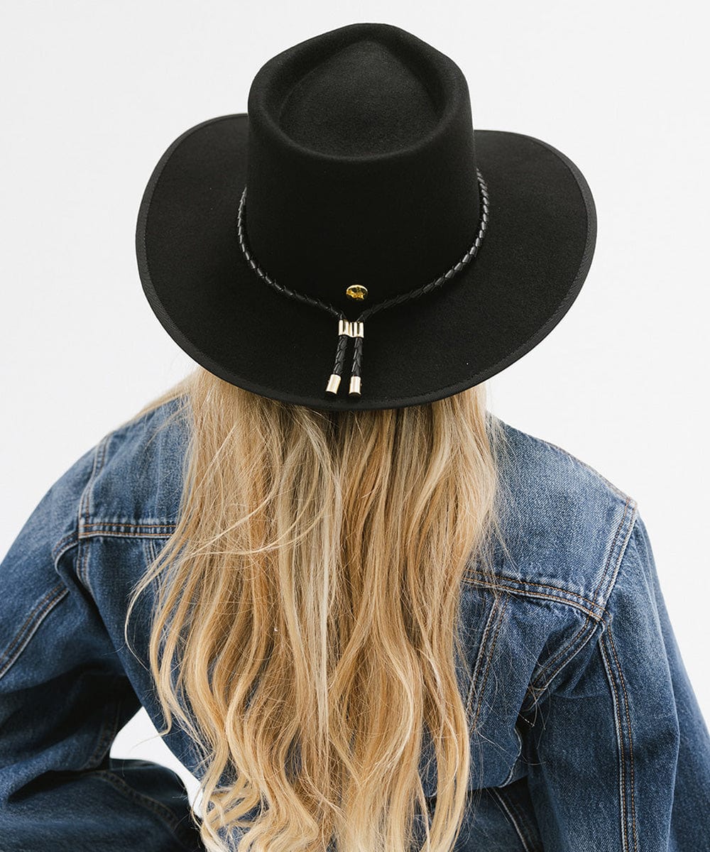 Woman with long hair wearing a black hat with a black braided bolo hat band and a denim jacket, facing behind against a plain background #color_black