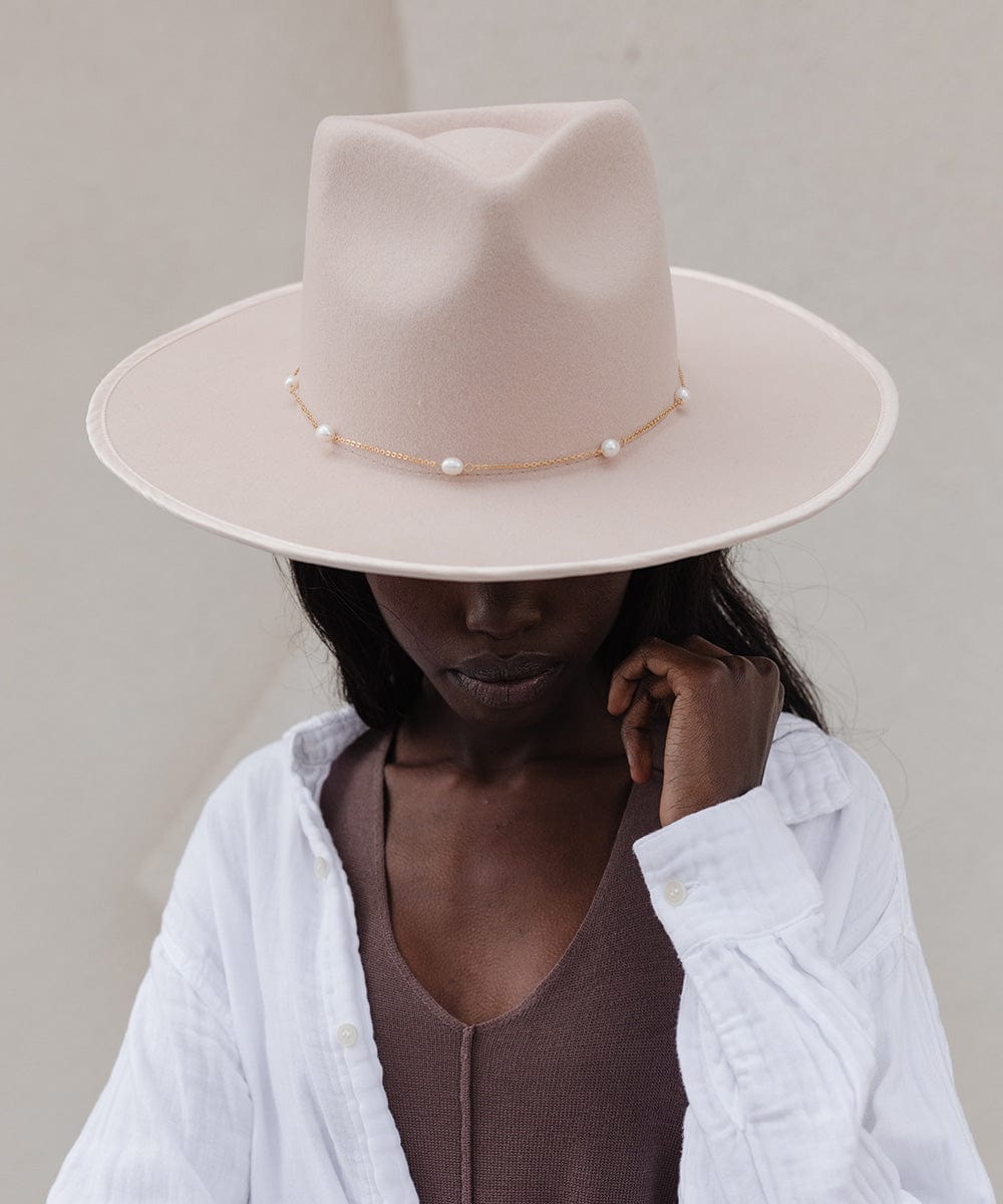 Woman wearing a cream hat with a gold pearl chain band and a brown top with a white shirt, looking down against a plain background #color_gold