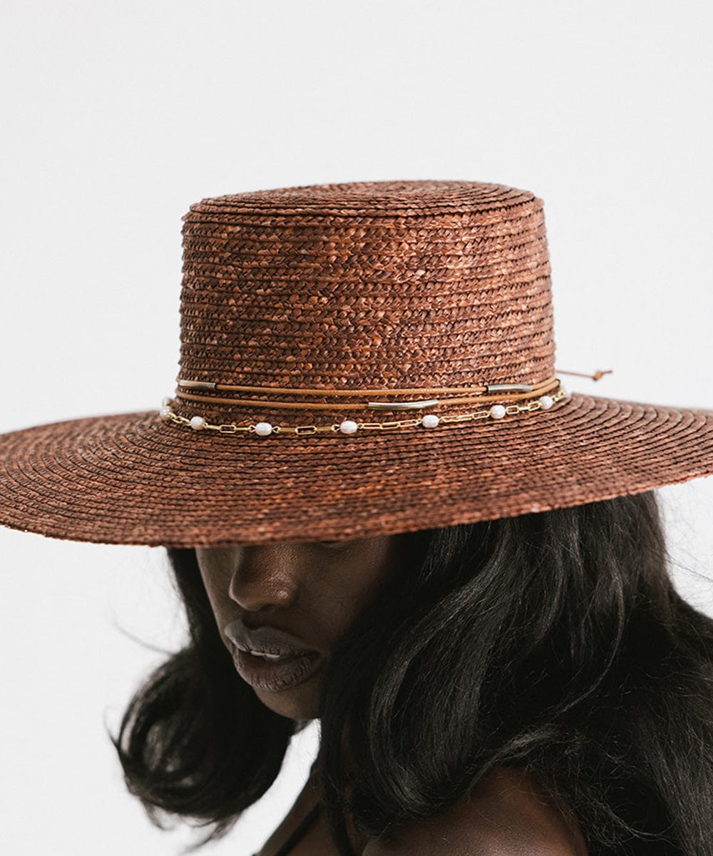 Woman wearing a brown straw hat with a gold pearl paperclip chain band against a plain background #color_gold
