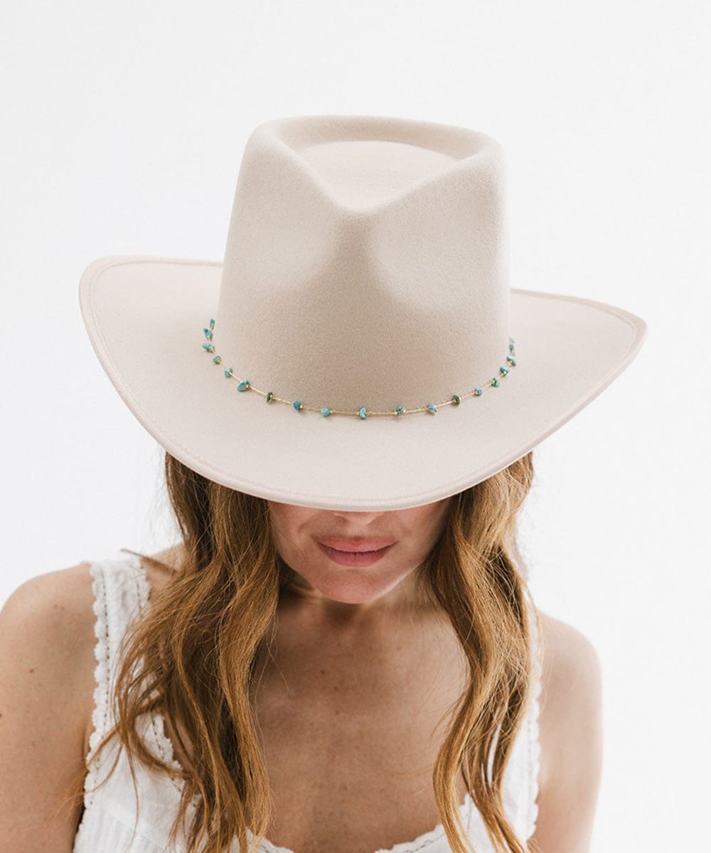 A woman wearing a cream hat with a turquoise stone hat band around the base and a white top against a plain background #color_gold