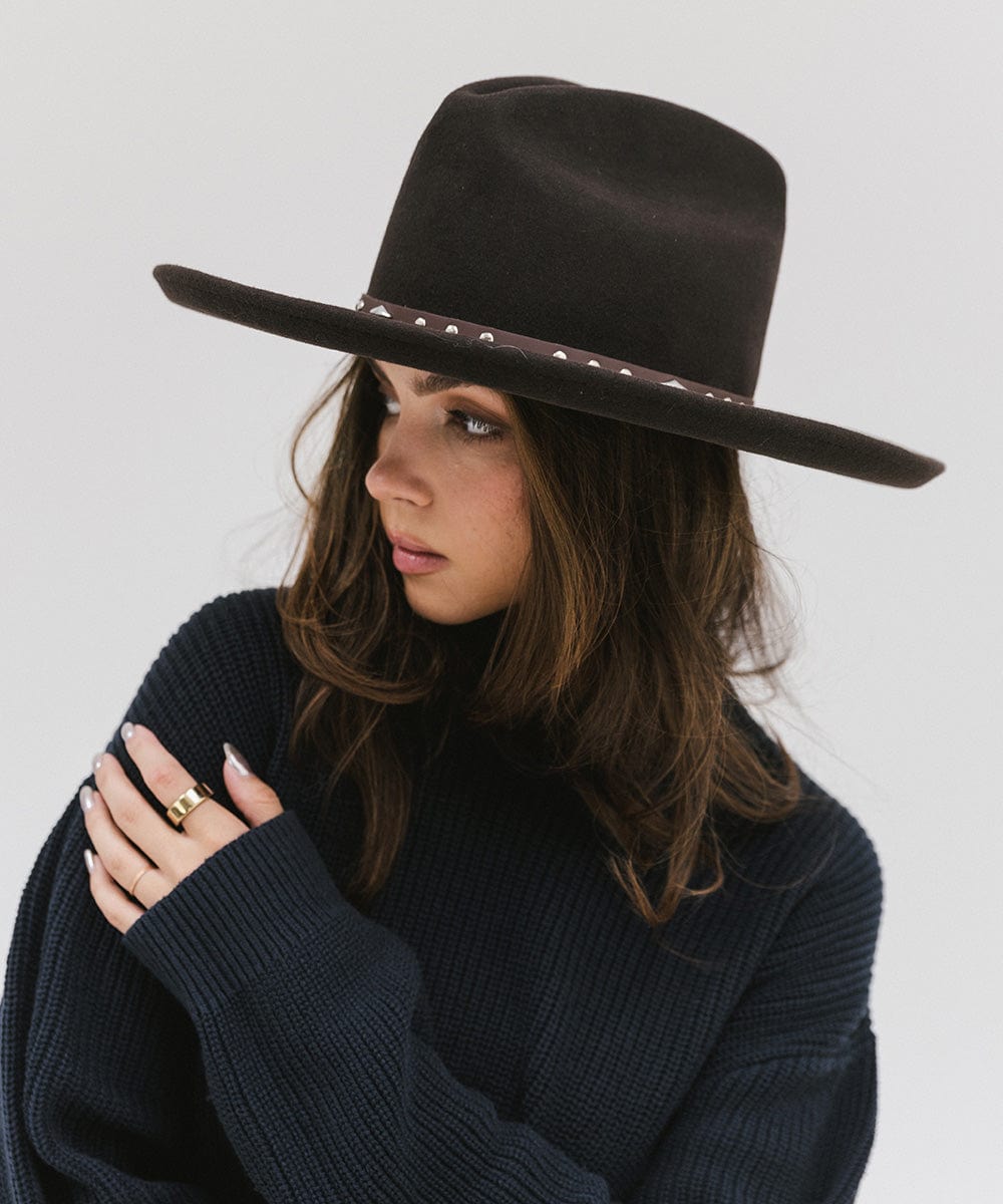 Woman wearing a brown hat with a chocolate colored studded leather hat band and a blue sweater, looking sideways against a plain background #color_chocolate