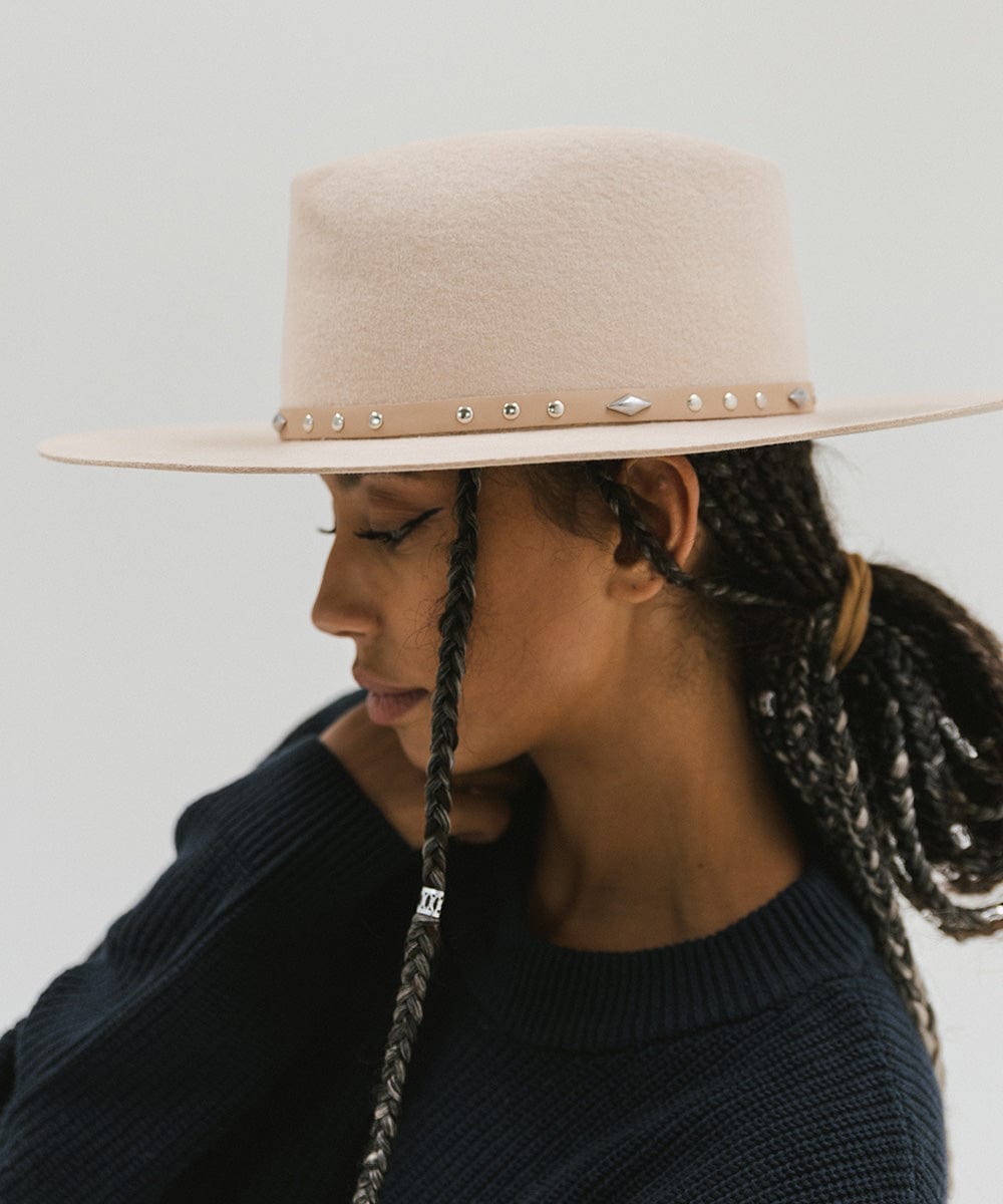 Woman wearing a cream hat with a tan studded leather hat band and a blue sweater, facing sideways against a plain background #color_tan