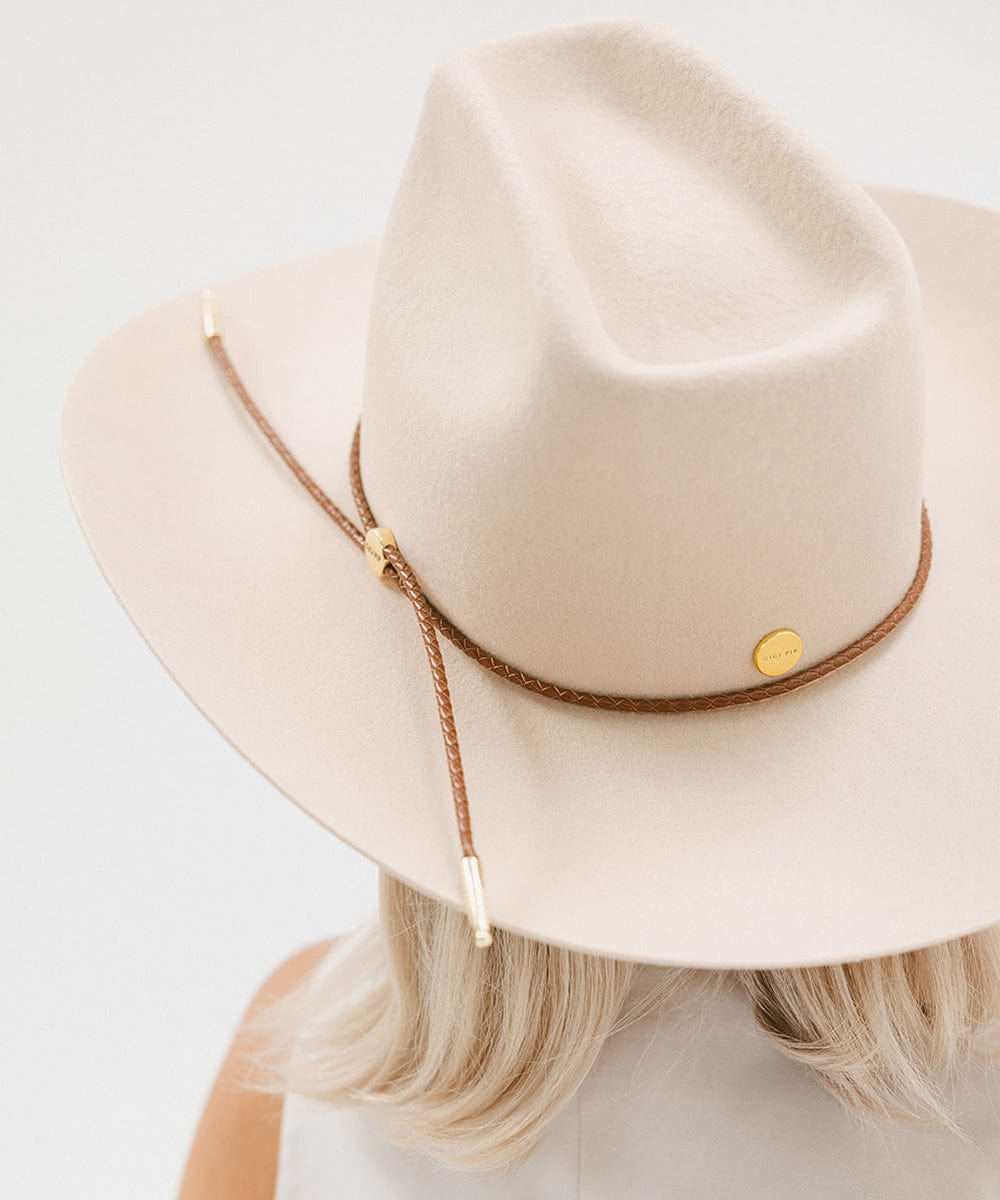 Woman wearing a cream cowboy hat with a cognac colored braided hat band and a white top, facing behind against a plain background #color_cognac