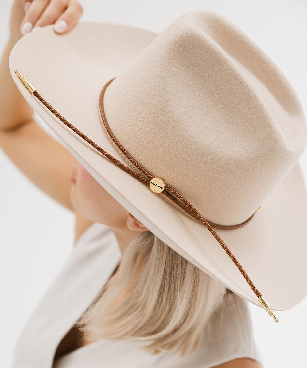 Woman wearing a cream cowboy hat with a cognac colored braided hat band and a white top, facing sideways against a plain background #color_cognac