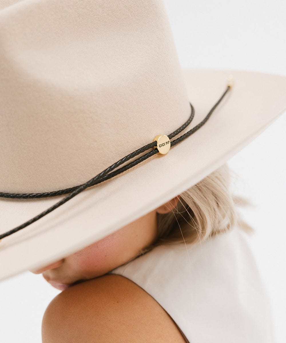 Close up image of a woman wearing a cream cowboy hat with a black braided hat band and a white top against a plain background #color_black