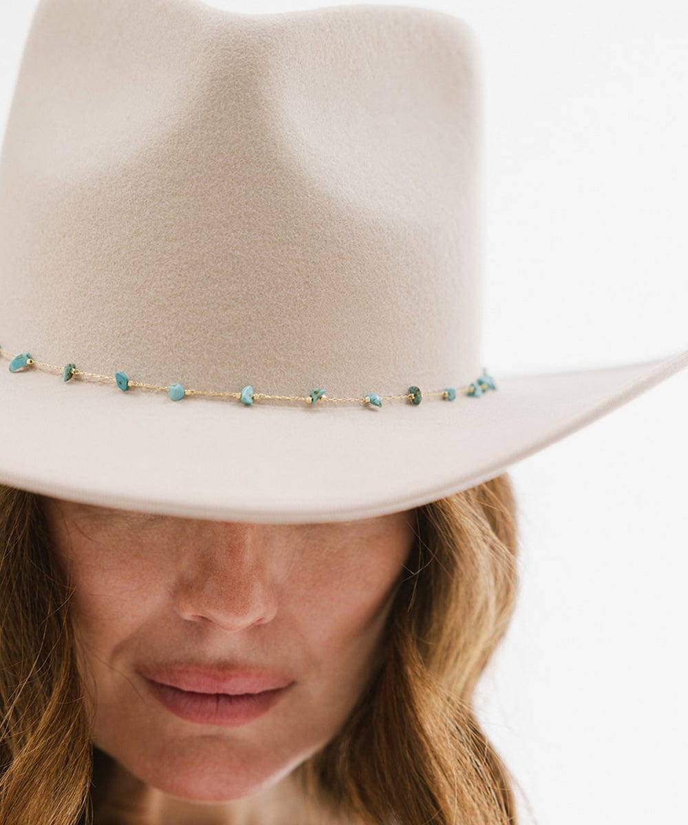 A woman wearing a cream hat with a turquoise stone hat band around the base against a plain background #color_gold