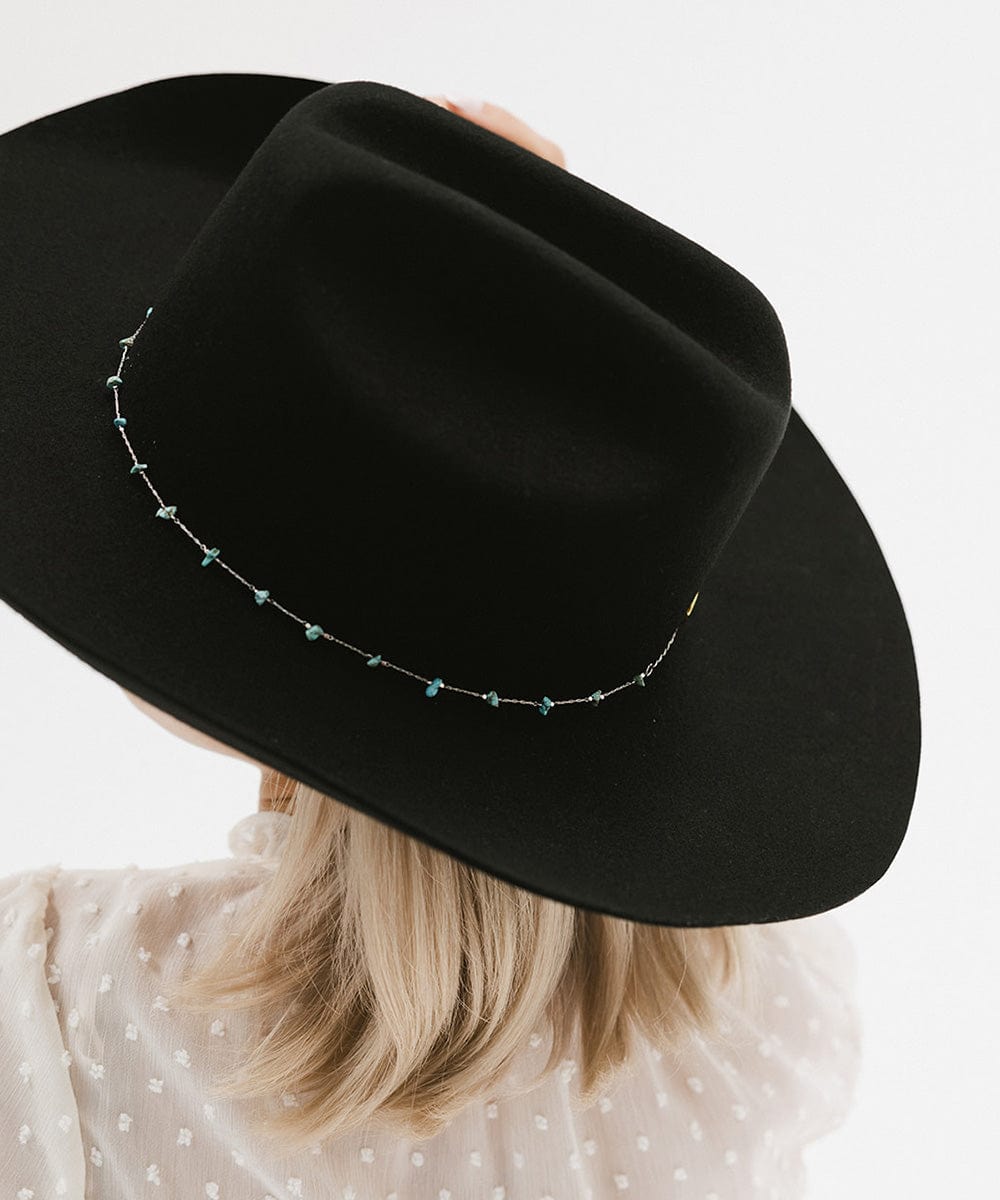 A person wearing a black hat with a turquoise stone hat band around the base and a white top, facing behind against a plain background #color_silver