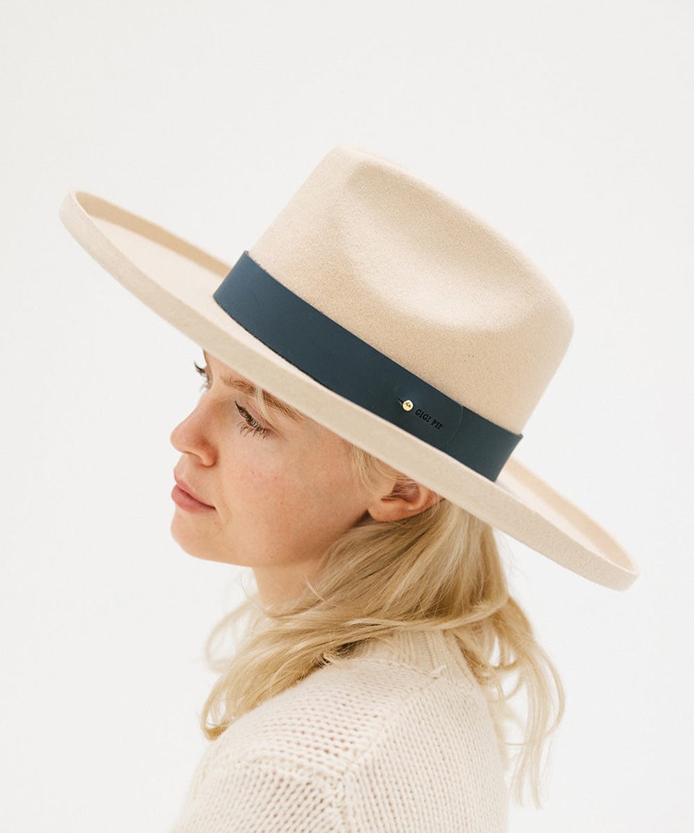 A woman wearing a cream hat with a wide vintage blue colored leather band around the base, facing sideways against a plain background #color_vintage blue