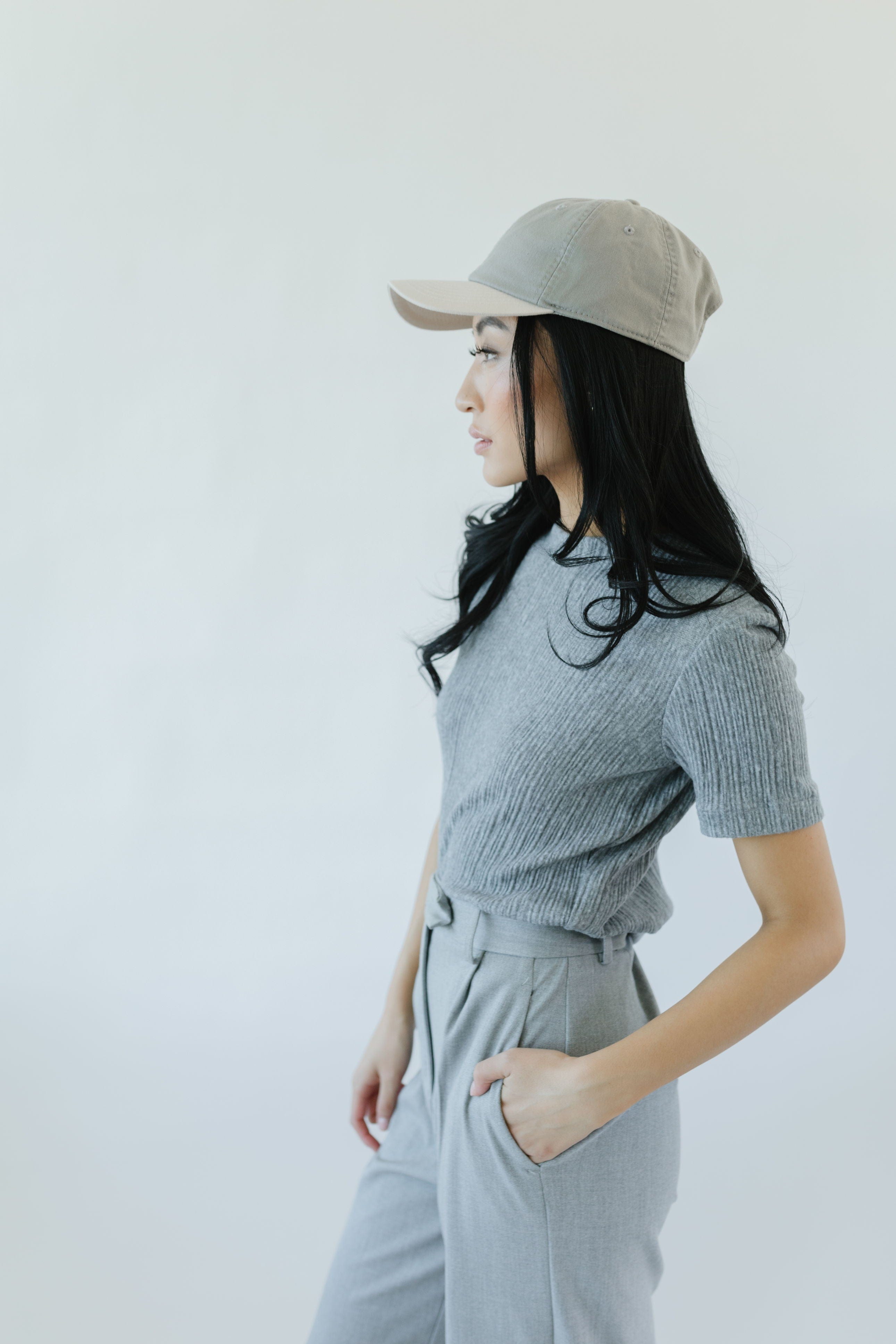 Woman wearing a light grey and cream colored ball cap, a blue t-shirt and grey pants, facing sideways against a plain background #color_light grey-cream