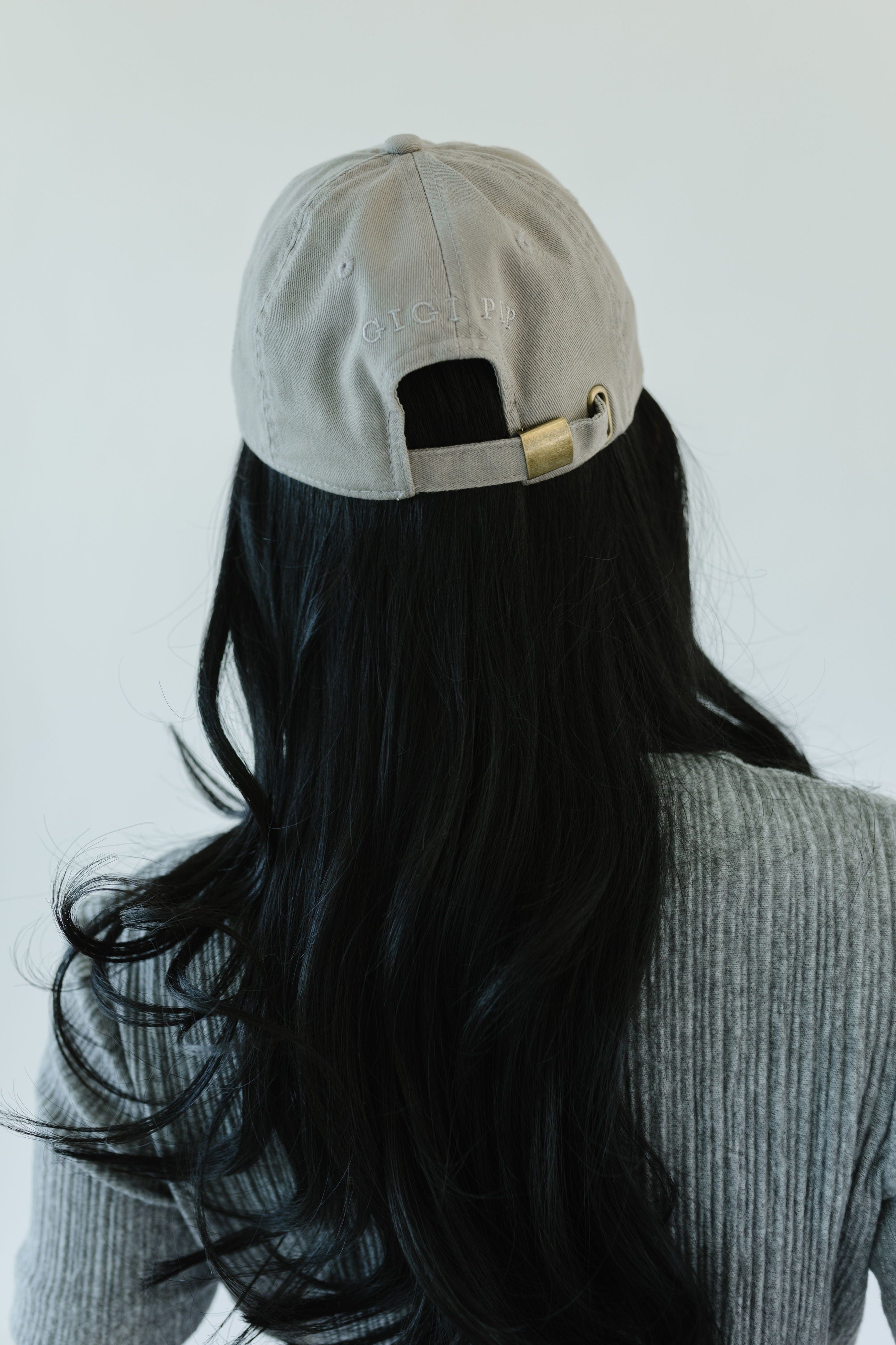 Woman wearing a light grey ball cap and a blue t-shirt, facing behind against a plain background #color_light grey-cream