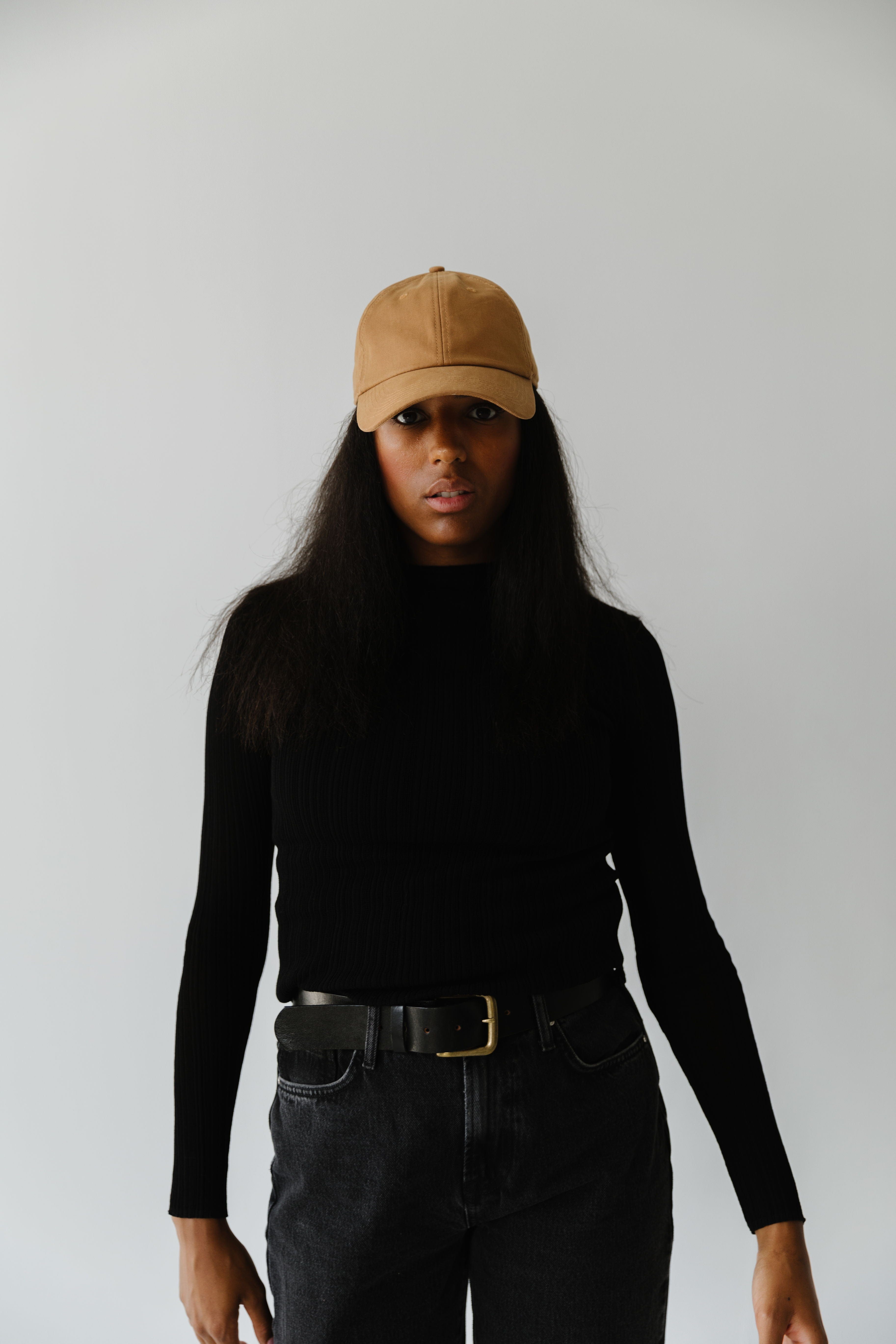 Woman wearing a camel colored ball cap, a black t-shirt and black jeans against a plain background #color_camel
