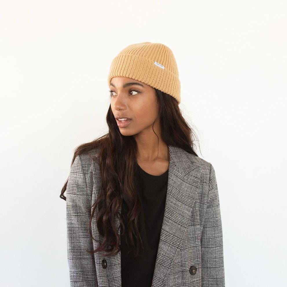 Woman wearing a camel colored knit beanie with the brand label and a black top with a blazer, looking sideways against a plain background #color_camel