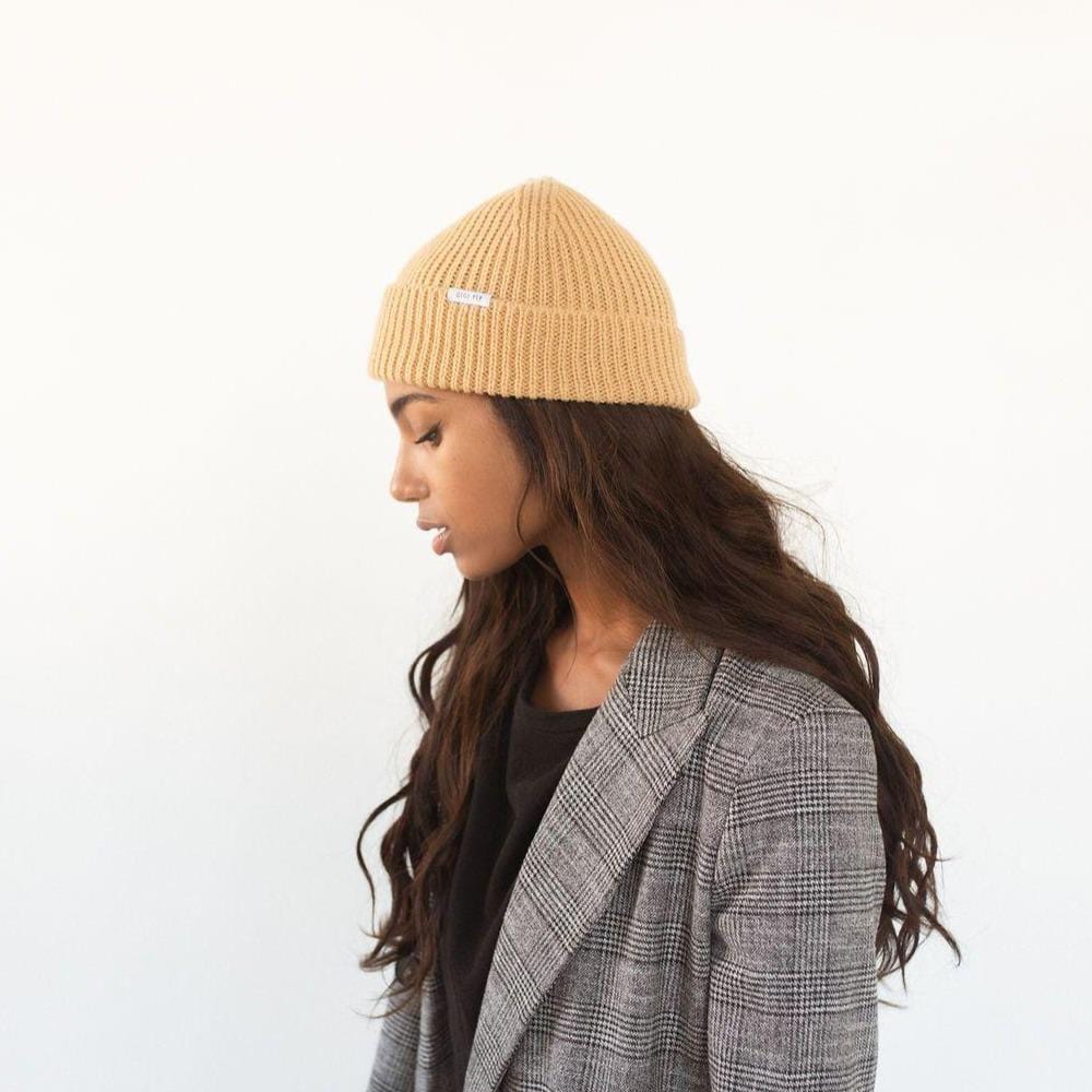 Woman wearing a camel colored knit beanie with the brand label and a black top with a blazer, facing sideways against a plain background #color_camel