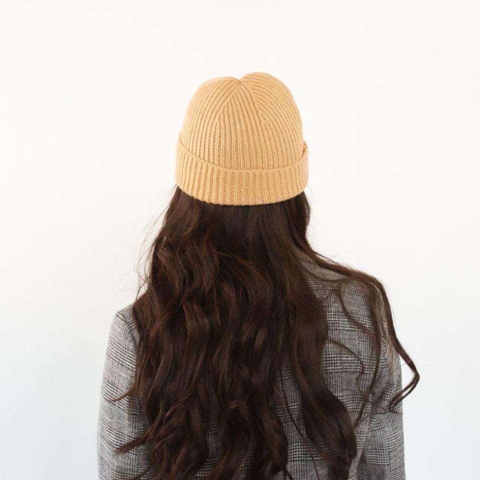 Woman wearing a camel colored knit beanie and a blazer, facing behind against a plain background #color_camel