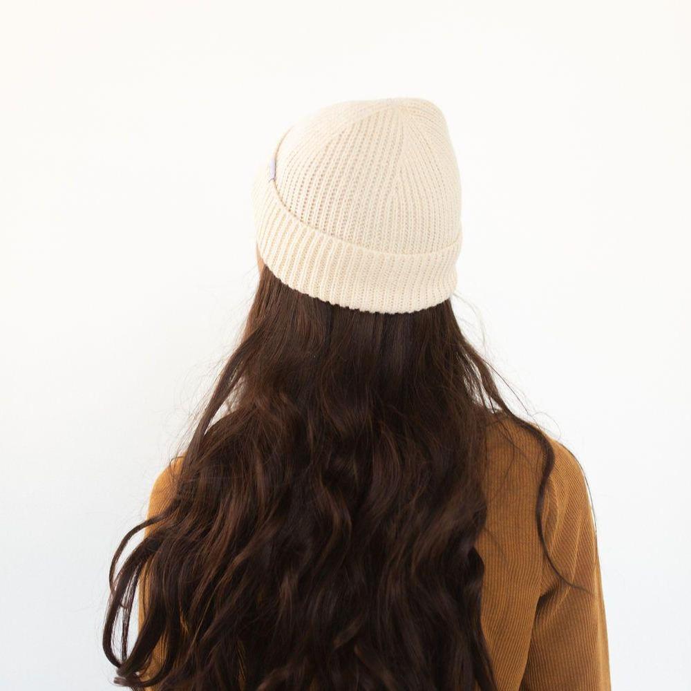 Woman wearing an oatmeal colored knit beanie and a brown sweatshirt, facing behind against a plain background #color_oatmeal