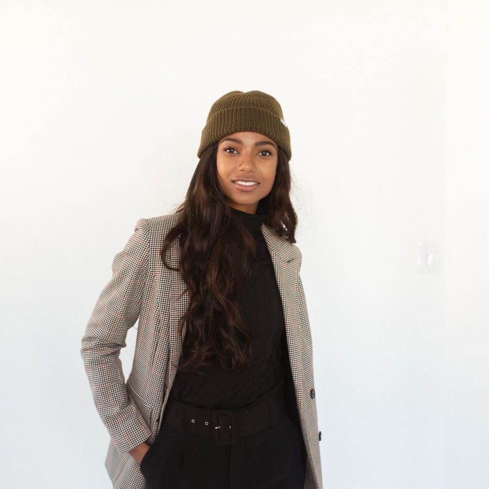 Woman wearing an olive colored knit beanie with the brand label, a black top with a blazer and black pants against a plain background #color_olive