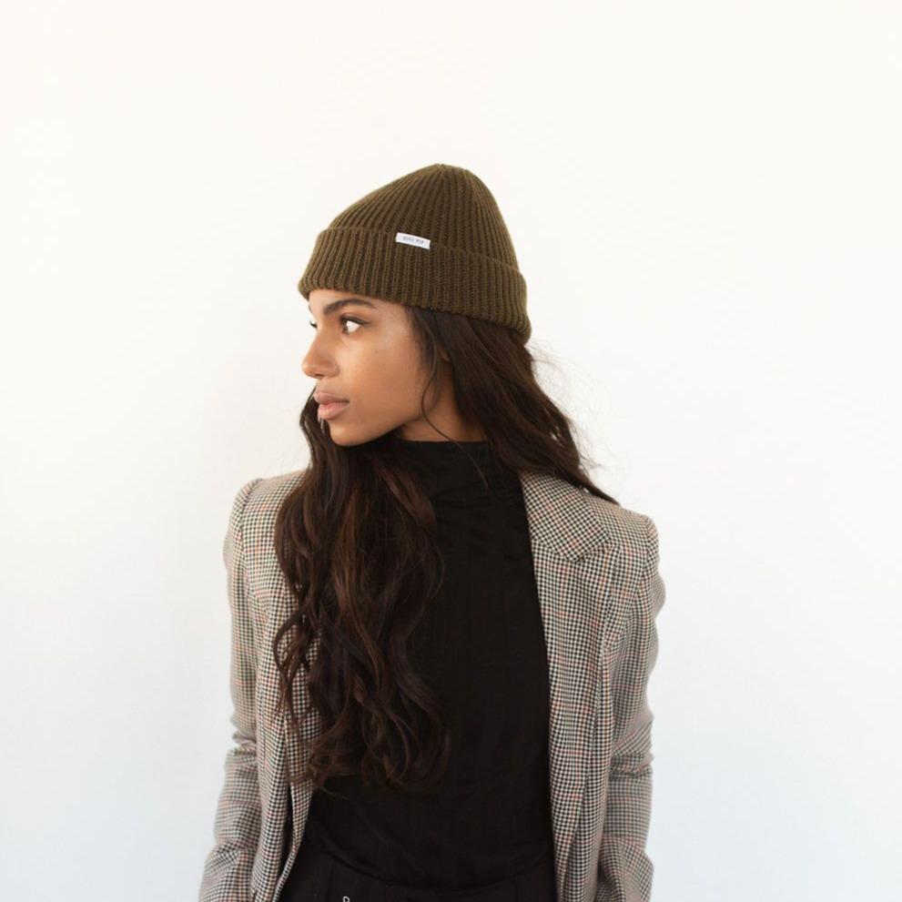 Woman wearing an olive colored knit beanie with the brand label and a black top with a blazer, looking sideways against a plain background #color_olive