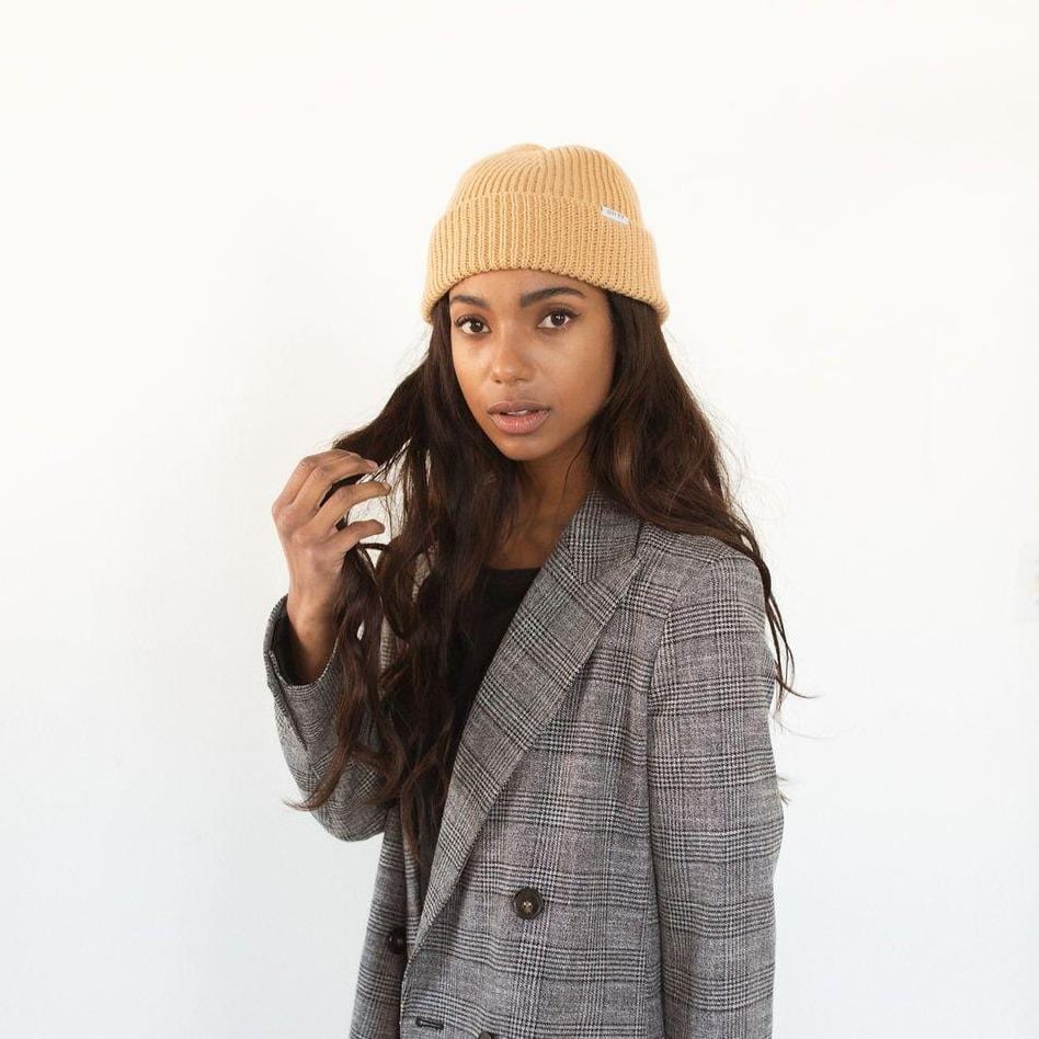 Woman wearing a camel colored knit beanie with the brand label and a black top with a blazer against a plain background #color_camel
