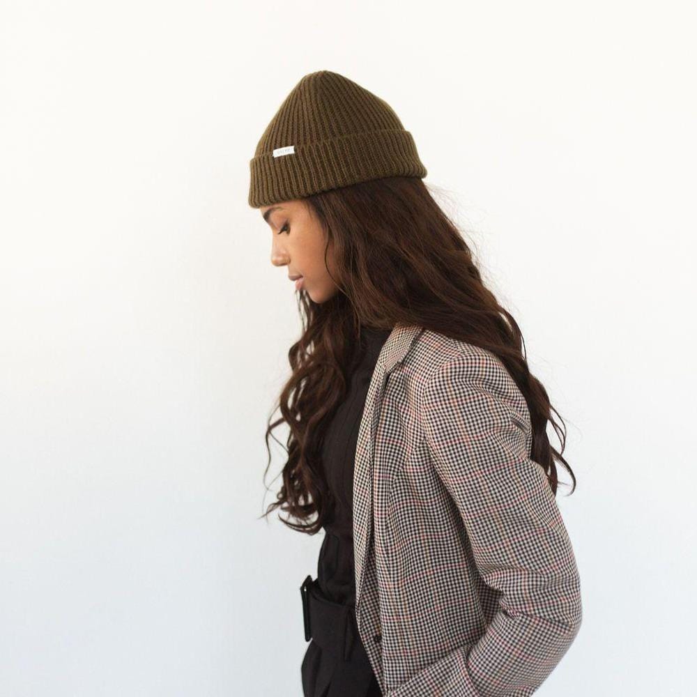 Woman wearing an olive colored knit beanie with the brand label, a black top with a blazer and black pants, facing sideways against a plain background #color_olive