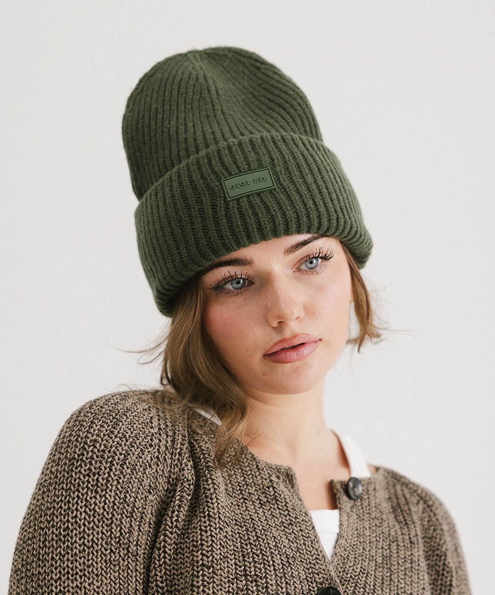 A woman wearing a dark green chunky ribbed beanie and a brown sweater against a plain background #color_dark green