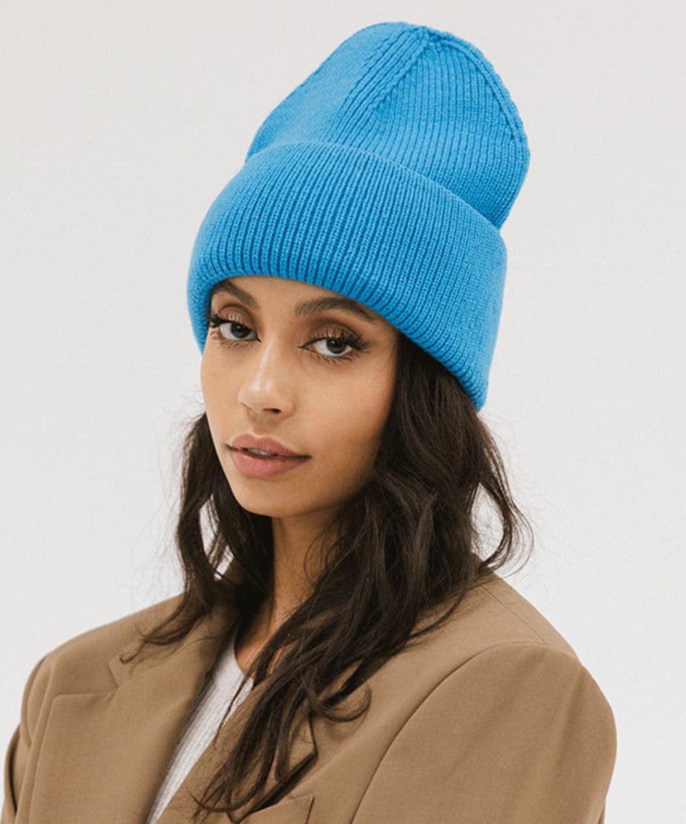 Woman wearing a blue oversized knit beanie and a brown coat against a plain background #color_deja blue