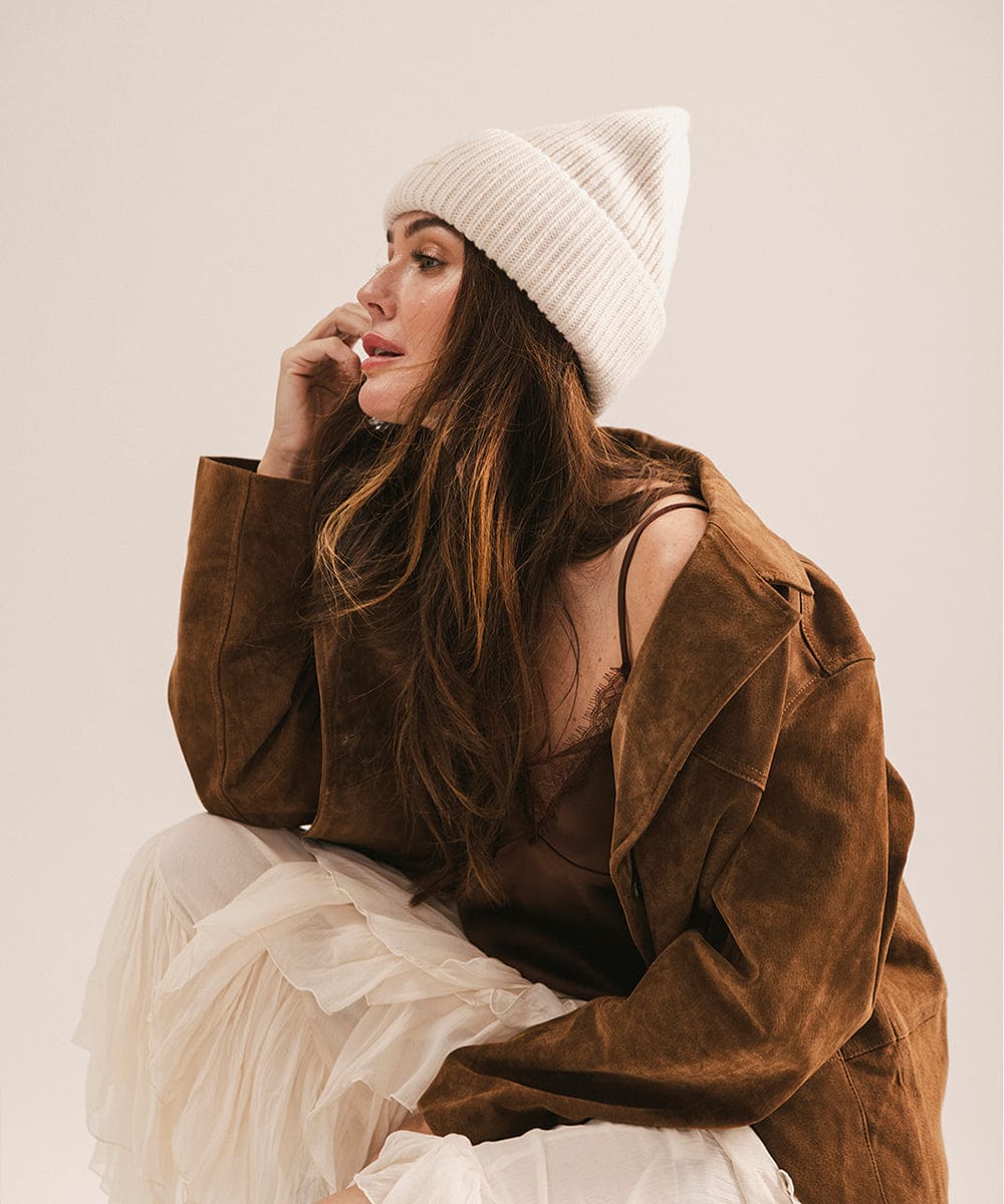 Woman wearing a brown jacket and white beanie sitting on a white surface. #color_off white