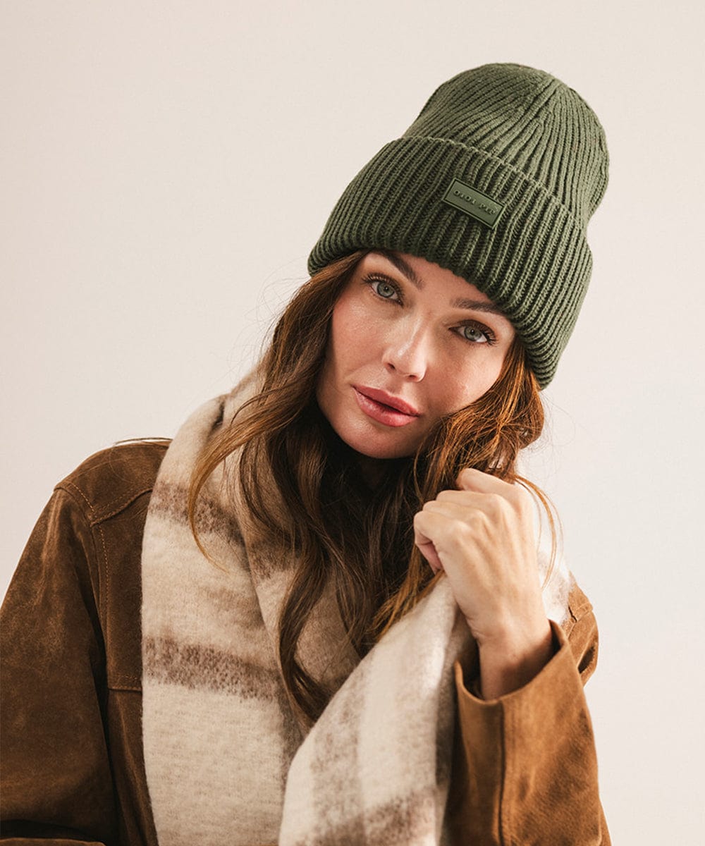 A woman wearing a dark green chunky ribbed beanie and a brown coat against a plain background #color_dark green