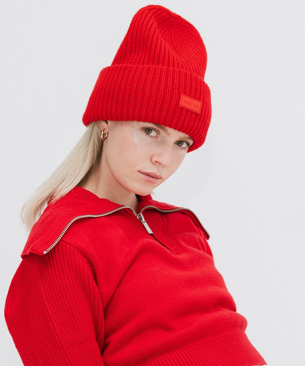 A woman wearing a ruby red colored ribbed beanie and a red jacket against a plain background #color_ruby red
